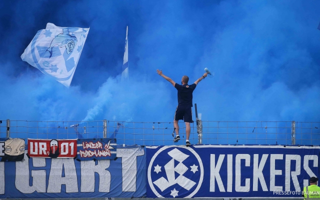 ein-Mann-steht-auf-einer-Mauer-welche-mit-Stuttgarter-Kickers-Bannern-voll-ist-der-Fanblock-ist-voll-mit-blauem-Nebel