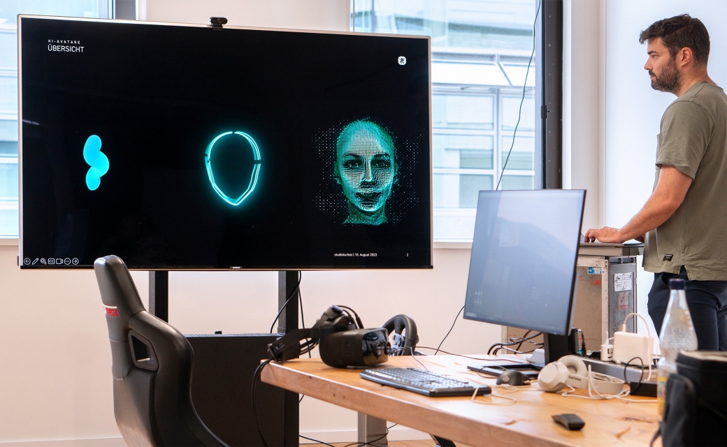 Ein-Mann-schaut-auf-einen-großen-Bildschirm-mit-drei-Versionen-von-Avataren-in-Mintgruen-davor-Kabel-Autositz-und-anderes-Equipment-zu-sehen