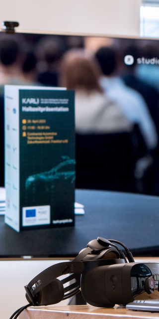 ein-bild-von-einem-großem-Bildschirm-mit-dem-Wort-Nutzerstudie-davor-ein-Autositz-mit-VR-Brille-auf-dem-Tisch